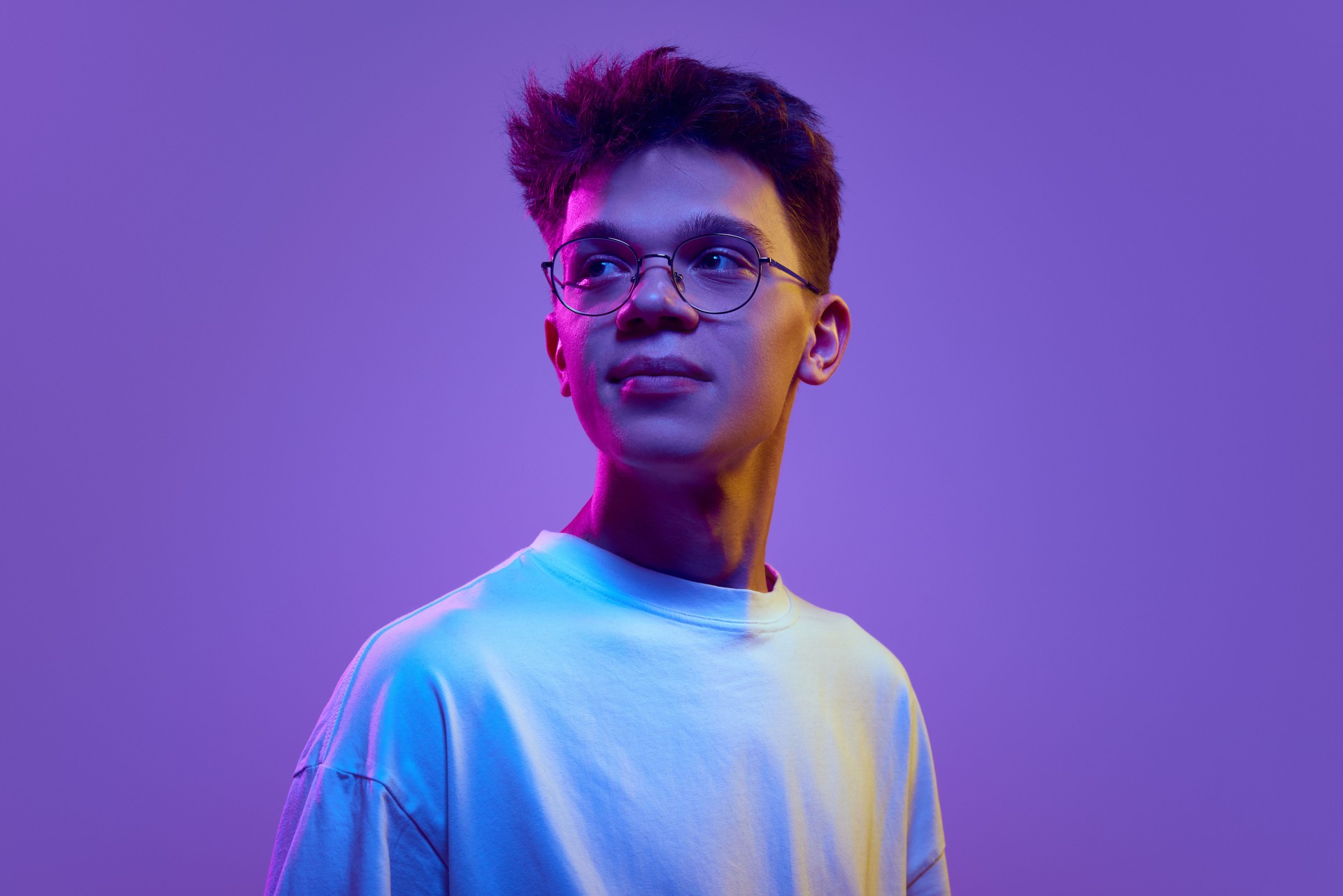 Portrait of smiling young man, boy in casual white t-shirt and sunglasses looking away against purple background in neon light. Relaxed look