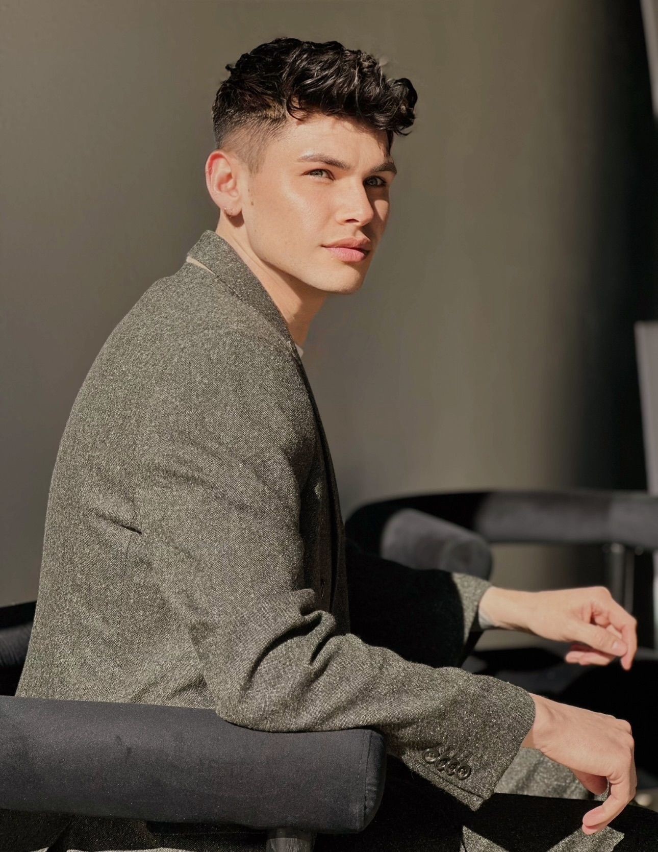 Man in a textured suit sitting on a chair, looking sideways with a thoughtful expression.